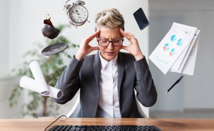 Image of a woman with hands to her temples with a phone, clock, spilling coffee cup and papers surrounding his head